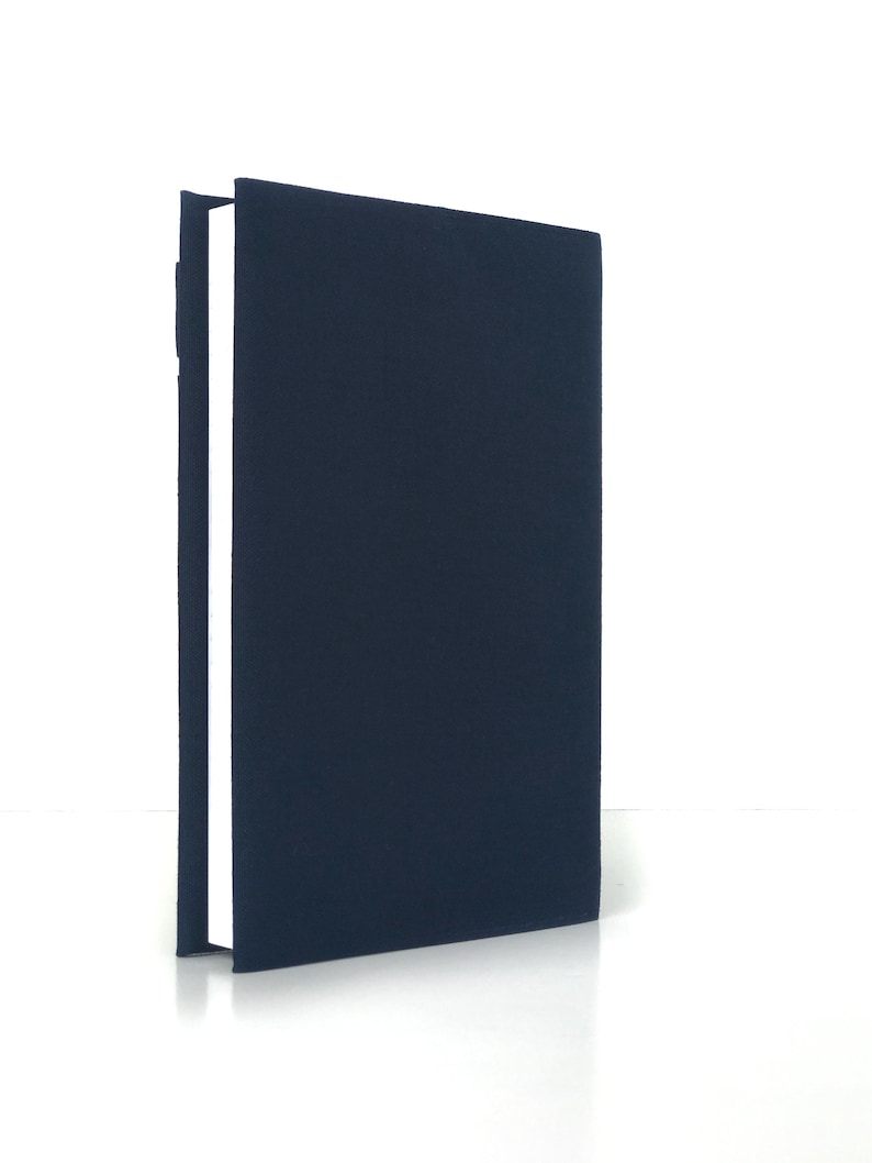 May include: A navy blue book with a textured cover stands upright against a white background. The book's spine is visible, revealing white pages. The cover is a solid, dark blue color.