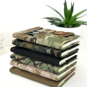 May include: A stack of six notebooks with covers in various camouflage patterns and a solid black cover. The top notebook has a pen holder with pens and patches. The notebooks are stacked on top of each other, with the pages visible.