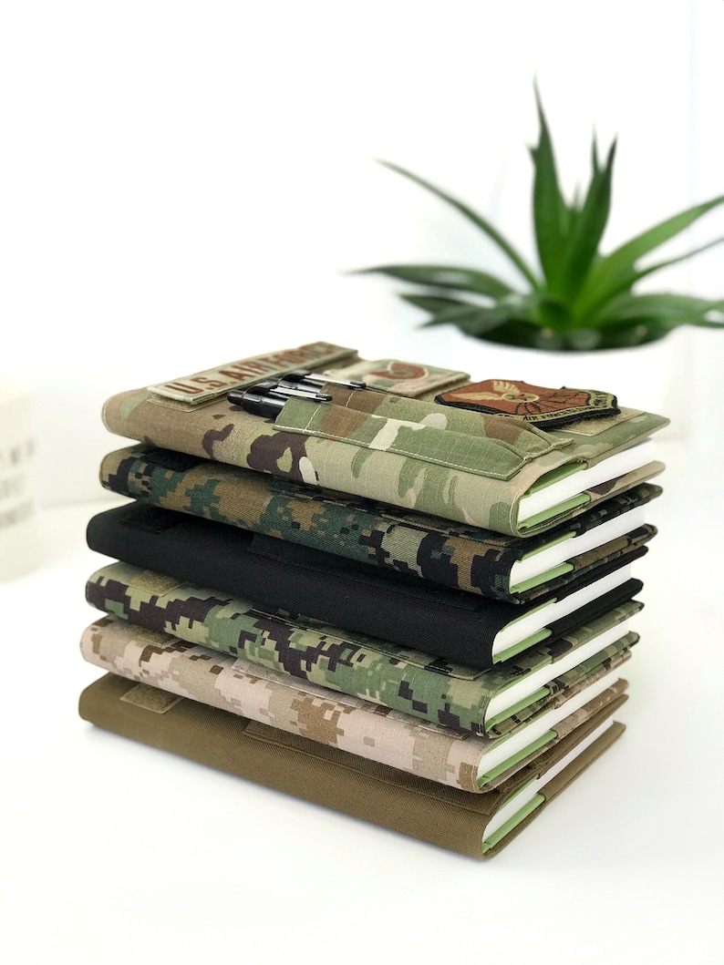 May include: A stack of six notebooks with covers in various camouflage patterns and a solid black cover. The top notebook has a pen holder with pens and patches. The notebooks are stacked on top of each other, with the pages visible.