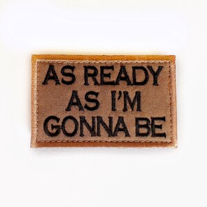 May include: A rectangular patch with the text "AS READY AS I'M GONNA BE" in black block letters. The patch has a brown background and a brown stitched border. The patch is centered on a white background.