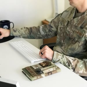 May include: A camouflage-patterned notebook cover with the text "U.S. AIR FORCE" and a patch. Two pens are in a pocket. A white keyboard and a black mug are also visible on the white desk.