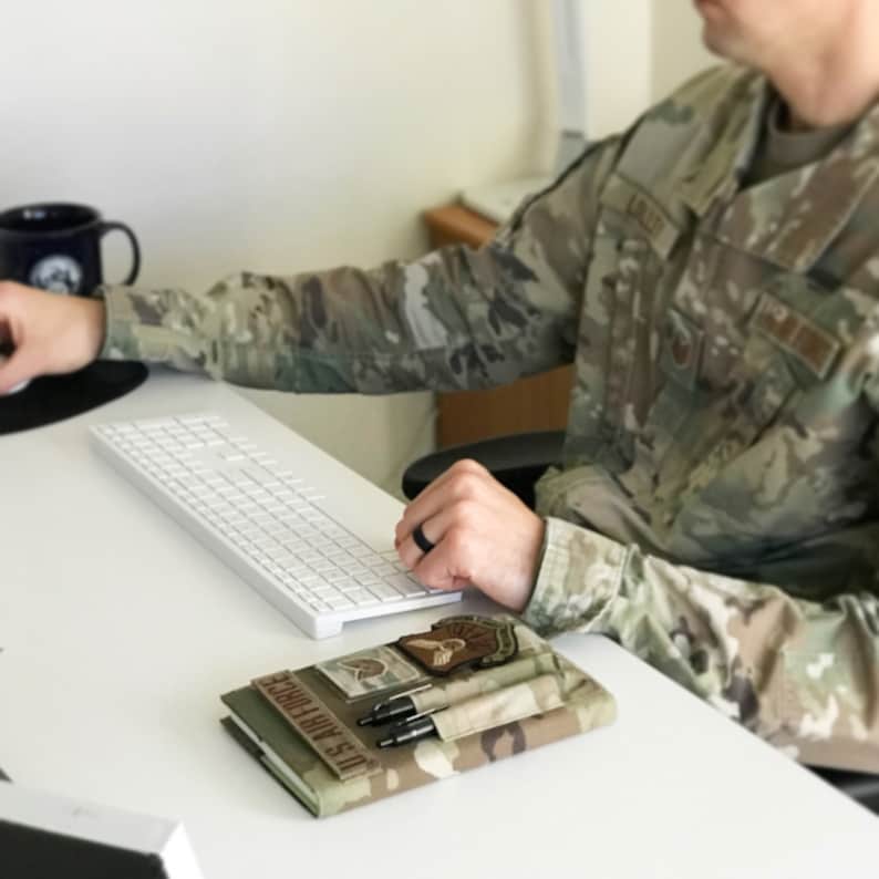 May include: A camouflage-patterned notebook cover with the text "U.S. AIR FORCE" and a patch. Two pens are in a pocket. A white keyboard and a black mug are also visible on the white desk.