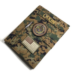 Pode incluir: Uma capa de caderno com padrão de camuflagem com o nome "LAKIN" bordado em dourado. Apresenta um patch do Corpo de Fuzileiros Navais dos Estados Unidos e um patch da bandeira americana. Uma caneta prateada está presa em um laço.