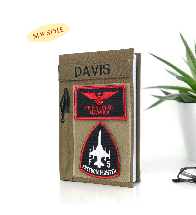 May include: A khaki-colored notebook cover with the name "DAVIS" embroidered on a top strip. It features a pen holder and two patches: one with a red eagle design and "PETE MITCHELL MAVERICK" text, and another with a jet and "FREEDOM FIGHTER" text. A "NEW STYLE" sticker is in the top left.