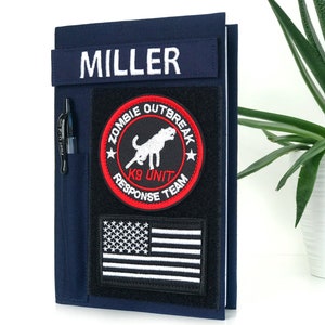 May include: Navy blue notebook cover with a black pen in a side pocket. The cover features a patch with the text "MILLER" and a "ZOMBIE OUTBREAK K9 UNIT RESPONSE TEAM" patch, along with an American flag patch.