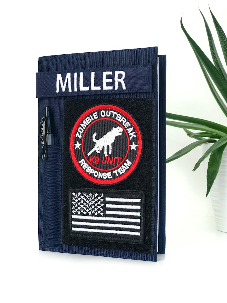 May include: Navy blue notebook cover with a black pen in a side pocket. The cover features a patch with the text "MILLER" and a "ZOMBIE OUTBREAK K9 UNIT RESPONSE TEAM" patch, along with an American flag patch.