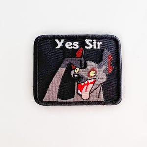 May include: A square, black embroidered patch with the text "Yes Sir" in white. The patch features a cartoon hyena with a gray body, red accents, and a red tongue sticking out. The hyena is smiling.