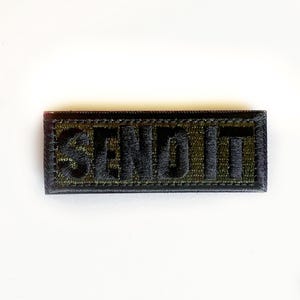 May include: Embroidered patch with the word "SEND IT" in black thread on a green and black background. The patch is rectangular and has a black border. The text is in bold, block letters.