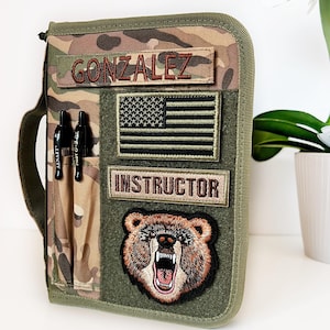 May include: Camouflage-patterned tactical notebook cover with the name "GONZALEZ" embroidered in brown. Features an American flag patch, the word "INSTRUCTOR", and a bear patch. Includes two black pens in a side pocket.