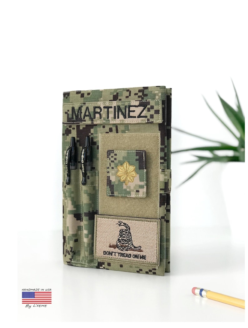May include: A camouflage-patterned notebook cover with the name "MARTINEZ" displayed on a patch. It features a pen holder, a military rank insignia, and a patch with the text "DON'T TREAD ON ME". The cover is handmade in the USA.
