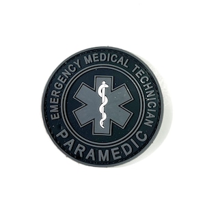 Peut inclure: Un écusson rond noir avec l'inscription "EMERGENCY MEDICAL TECHNICIAN PARAMEDIC" encerclant un symbole gris en forme d'étoile de vie. Le symbole comprend un bâton blanc avec un serpent.