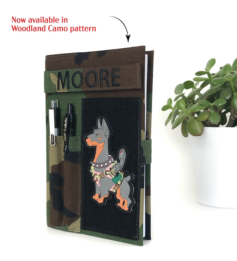 May include: Woodland camo pattern notebook cover with a black patch featuring a cartoon Doberman Pinscher. The cover has a pen holder and a name patch that says "MOORE". The text "Now available in Woodland Camo pattern" is also visible.