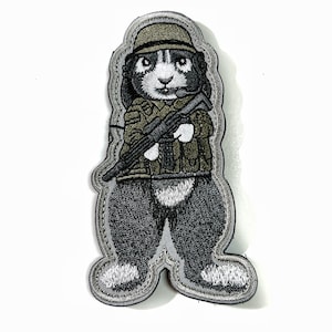 Peut inclure: Écusson brodé représentant un lapin en uniforme militaire, tenant un fusil. Le patch a une bordure grise. Le lapin est représenté en uniforme et casque vert, avec des détails noirs, blancs et gris.