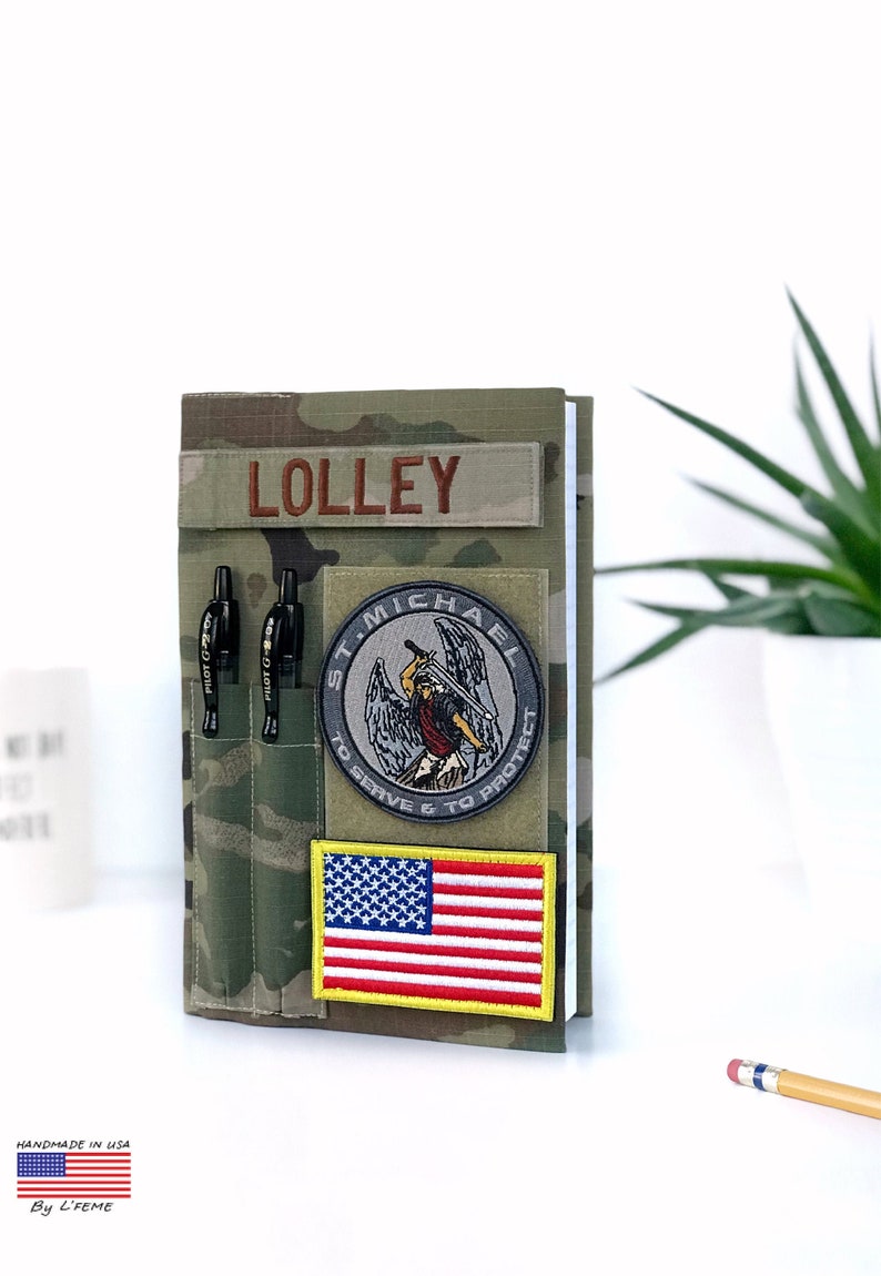 May include: A camouflage-patterned notebook cover with the name "LOLLEY" embroidered in brown. It features patches including the American flag and a St. Michael emblem. Two black pens are in a pocket. "HANDMADE IN USA" is printed at the bottom.