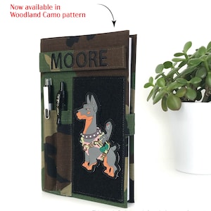 May include: Woodland camo pattern notebook cover with the name "MOORE" embroidered on a fabric strip. The cover features a black patch with a cartoon dog design and pen holders. The text "Now available in Woodland Camo pattern" is visible.