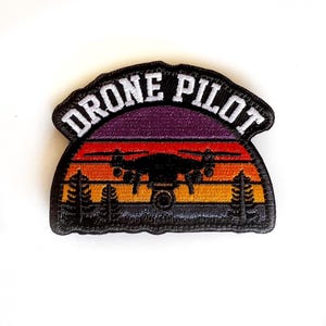 May include: Embroidered patch with the words "DRONE PILOT" in white arched over a sunset scene. The sunset features shades of purple, red, and orange, with a black drone silhouette and trees.