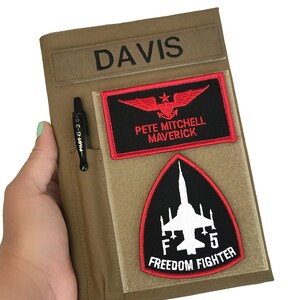 May include: Tan notebook cover with the name "DAVIS" embroidered in black. It features a pen holder with a black pen and two patches: one with red wings and the text "PETE MITCHELL MAVERICK", and another with a jet and the text "F5 FREEDOM FIGHTER".