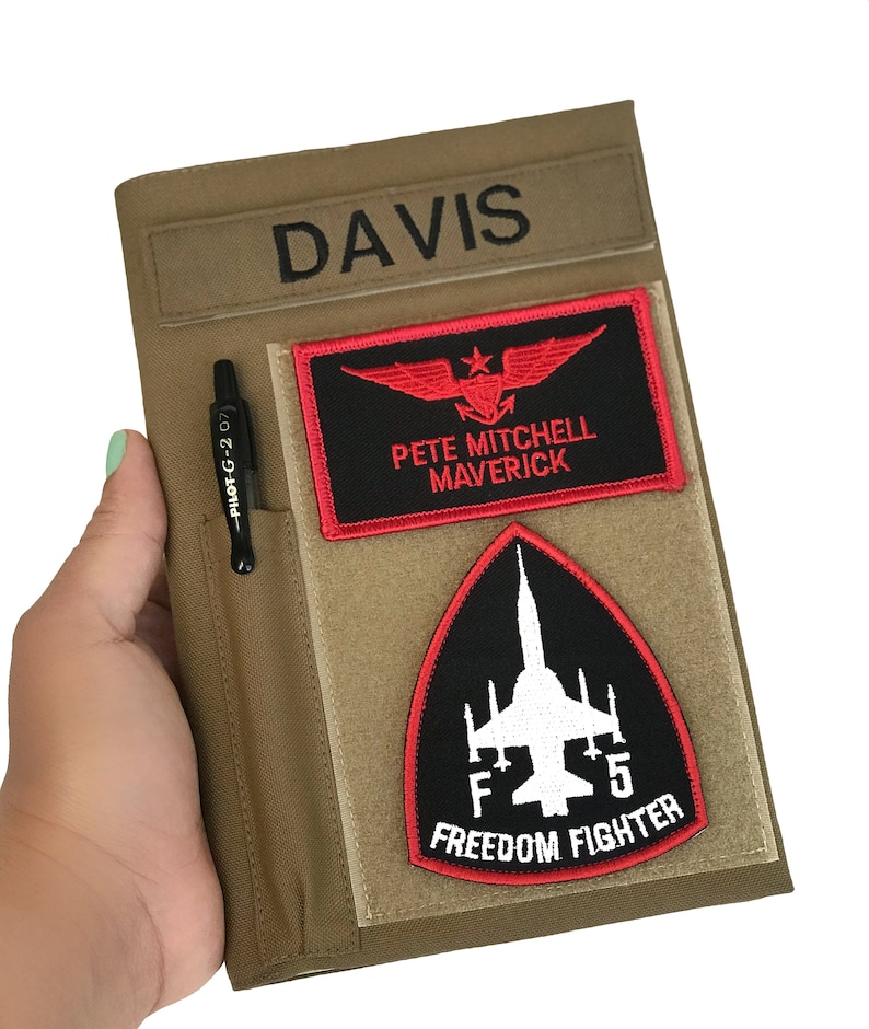 May include: Tan notebook cover with the name "DAVIS" embroidered in black. It features a pen holder with a black pen and two patches: one with red wings and the text "PETE MITCHELL MAVERICK", and another with a jet and the text "F5 FREEDOM FIGHTER".