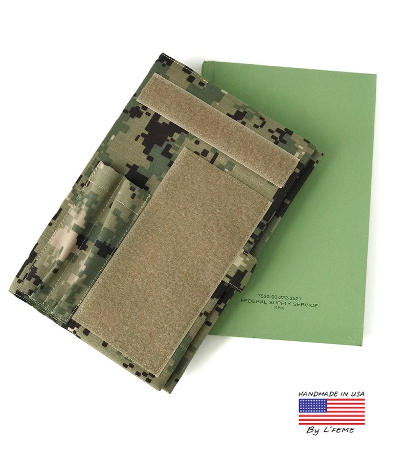 May include: A camouflage pattern notebook cover with hook and loop fasteners, next to a green notebook. The cover is made of a digital camouflage fabric. The text "7530-00-222-3521 FEDERAL SUPPLY SERVICE (FSC)" is visible on the green notebook. "HANDMADE IN USA" is also visible.