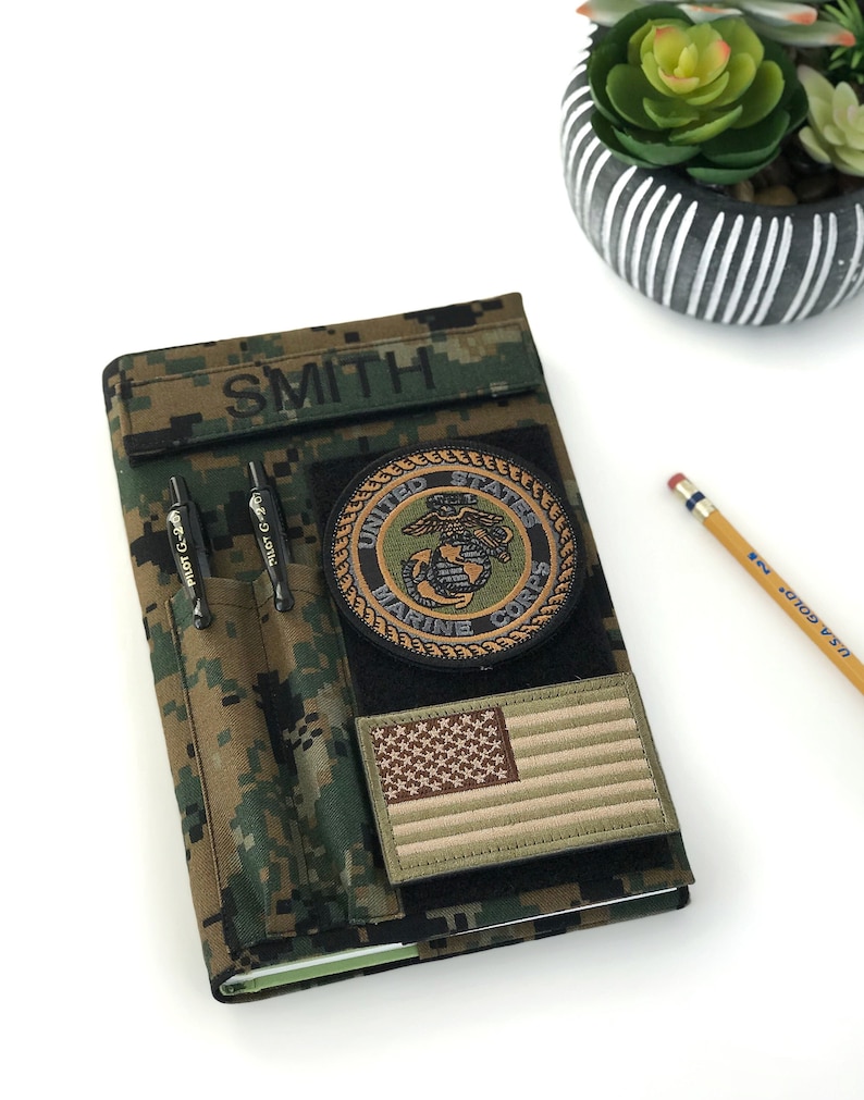 May include: A camouflage-patterned notebook cover with the name "SMITH" above two pen holders. It features a "United States Marine Corps" patch, an American flag patch, and two black pens. A pencil and a decorative plant are in the background.