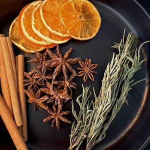 May include: A black plate containing dried orange slices, several brown cinnamon sticks, a cluster of star anise pods, and a sprig of dried green rosemary. The ingredients are arranged on the dark surface of the plate for display.