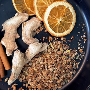 May include: A black plate containing three dried orange slices, three pieces of raw ginger root, two cinnamon sticks, and a pile of small, brown, granular herbal tea ingredients scattered across the surface of the plate.