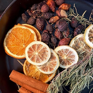 May include: A black bowl containing dried orange slices, dried lemon slices, cinnamon sticks, sprigs of green rosemary, and dark brown rose hips. The arrangement of dried fruits, herbs, and spices is displayed together in the round container.