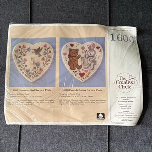 May include: A Creative Circle cross-stitch kit package containing two heart-shaped switch plate designs: 1601 Hummingbird and 1606 Bear & Bunny. The kit includes 14-count Aida cloth, cotton embroidery floss, a #24 tapestry needle, and an acrylic cover.