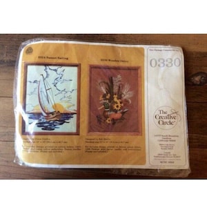 Puede incluir: Un kit de punto de cruz empaquetado con dos diseños enmarcados: "Sunset Sailing" con un velero y "Wooden Decor" con un arreglo floral y un pato. El kit incluye tela estampada, hilo, agujas e instrucciones.