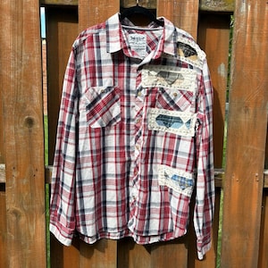 May include: A long-sleeve button-down shirt featuring a red, white, and navy blue plaid pattern. The shirt includes a collar, a front button placket, and several rectangular fabric patches with visible cream-colored stitching on the front.
