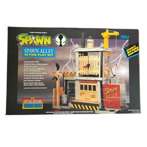 Op de afbeelding: Todd McFarlane's Spawn Alley actiespeelset in een doos. De set bevat een structuur met meerdere niveaus, een kraan, een rode bakstenen muur met het opschrift Spawn Alley, een garage en een telefoonpaal. De verpakking bevat tekst met details over functies zoals een exploderende muur, een raketwerper en een stripboek.