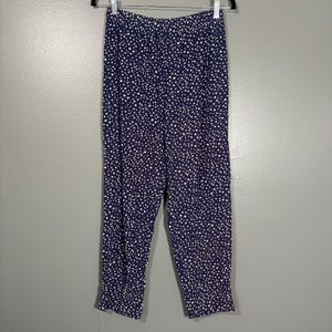 May include: Navy blue trousers with a white polka dot pattern. The trousers are hanging on a metal hanger against a grey background. The trousers have an elasticated waistband and gathered cuffs at the ankles.