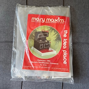 May include: A Mary Maxim needlecraft kit in a clear plastic bag containing a plastic canvas sheet and a red instruction sheet. The red sheet features a graphic of a black stove ornament, the text Christmas 1985 Collectors Needlepoint Ornament, and the idea place.