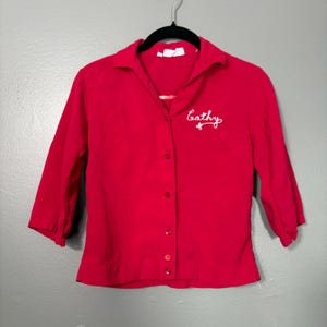 May include: A bright red button-down shirt with three-quarter sleeves. The shirt has a collar and is embroidered with the name "Cathy" in white script on the left chest. The shirt is hanging on a black hanger against a gray background.