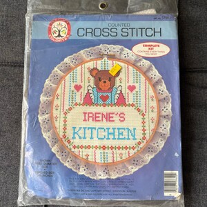 Op de afbeelding: Een Colortex telpatroon kruissteekpakket in een doorzichtige plastic zak. De verpakking toont een cirkelvormig ontwerp met een bruine beer, roze hartjes en de tekst 'IRENE'S KITCHEN' in roze en blauw. Het ontwerp is omrand met witte broderie.