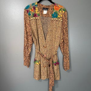 May include: A vintage button-down cardigan with a floral design. The long-sleeved cardigan features a brown and tan speckled pattern, a matching belt, and colorful floral accents on the shoulders and pockets. The cardigan is hanging on a black hanger.