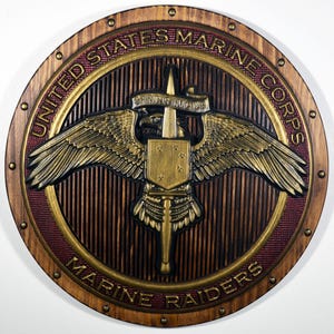 以下が含まれることがあります： アメリカ海兵隊のエンブレムが描かれた木製の円形プラーク。デザインには、金色の文字で「United States Marine Corps」と「Marine Raiders」の文字が含まれています。エンブレムは、金色のワシ、盾、剣、そしてモットー「Spiritus Invictus」が特徴です。