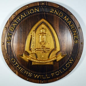 May include: A round wooden plaque with the text "1st Battalion 2nd Marines" and "OTHERS WILL FOLLOW" in gold lettering. The center features a gold emblem with the number "2" and other military symbols. The wood has a dark brown stain.