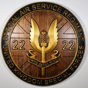 Op de afbeelding: Ronde houten plaquette met het embleem van het Special Air Service Regiment. Het ontwerp omvat een gouden zwaard en vleugels, het nummer "22" en het motto "WHO DARES WINS". De tekst "UNITED KINGDOM SPECIAL FORCES" en "SPECIAL AIR SERVICE REGIMENT" is ook aanwezig.