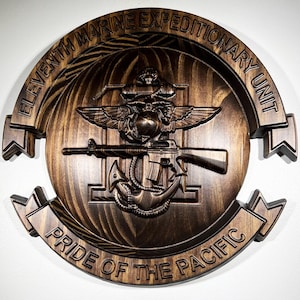 May include: A wooden plaque with the text "ELEVENTH MARINE EXPEDITIONARY UNIT" and "PRIDE OF THE PACIFIC". The plaque features a detailed relief of a military emblem with an eagle, globe, anchor, and rifle. The wood has a dark brown finish.
