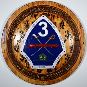 May include: A wooden plaque with a circular design featuring the 3rd Recruit Training Battalion logo. The logo is a blue shield with a white number 3, crossed swords, and the words "Spirit" and "Discipline" on a red ribbon. The shield is surrounded by a gold border with the words "3rd Recruit Training Battalion" and "We Make Marines".