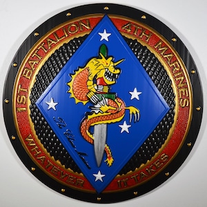 May include: A round, decorative emblem featuring the text "1st Battalion 4th Marines" and "Whatever It Takes." The emblem has a blue diamond with a yellow dragon and a dagger. The outer ring is black and red.