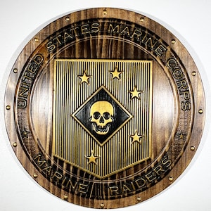 Op de afbeelding: Ronde houten plaquette met de tekst "UNITED STATES MARINE CORPS MARINE RAIDERS" gegraveerd langs de rand. Het midden toont een goud en zwart embleem met een schedel en sterren.