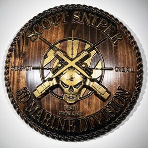 May include: A circular wooden wall plaque featuring a gold-toned relief of a skull and crossed rifles. The text reads SCOUT SNIPER, ONE SHOT ONE KILL, 3D MARINE DIVISION, and DEATH FROM AFAR. The border is carved with a rope-like design.