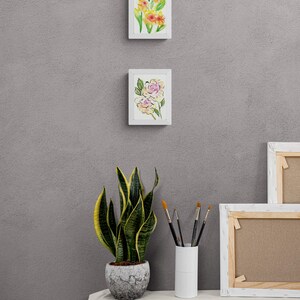 May include: Two framed watercolor paintings of flowers, one with yellow flowers and one with pink flowers, hanging on a gray wall. A potted snake plant and a vase of paintbrushes are on a white table in front of the wall.