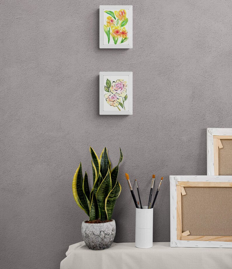 May include: Two framed watercolor paintings of flowers, one with yellow flowers and one with pink flowers, hanging on a gray wall. A potted snake plant and a vase of paintbrushes are on a white table in front of the wall.