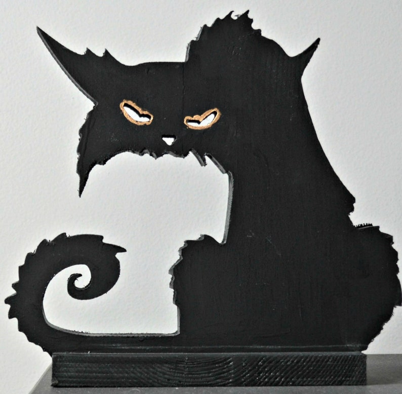 Op de afbeelding: Zwarte houten silhouet van een kat met een gekrulde staart. De kat heeft een dreigende uitdrukking met grote, witte ogen.