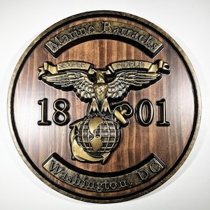 Può includere: Una targa da parete circolare in legno con l'emblema della Marine Barracks in finitura oro metallizzato. Il design include un'aquila, un globo e un'ancora, con il testo Marine Barracks, Semper Fidelis, 1801 e Washington, DC su venature del legno.