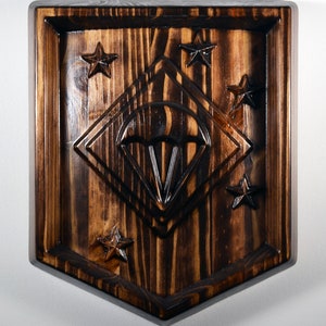 May include: A wooden plaque with a carved image of a parachute inside a diamond shape. The plaque is surrounded by five stars and has a dark brown wood finish.