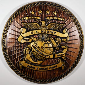 May include: A circular wooden plaque featuring a carved relief of the U.S. Marine Corps emblem. The emblem includes an eagle, globe, and anchor, with the text "U.S. MARINE" and "Semper Fidelis". The plaque is brown with gold detailing.